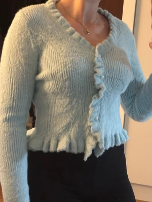 Ljusblå fluffig kofta med volang - Supersöt ljusblå kofta i fluffigt material med volangkant längs nederdelen och knäppning framtill. Koftan har långa ärmar och en figurnära passform som ger en trendig look. Passar xs-S, aldrig använd.