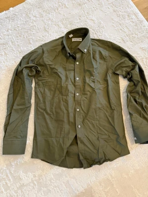 Olivgrön skjorta från Lacoste - Snygg olivgrön skjorta från Lacoste med klassisk button-down krage och vita knappar. Skjortan har lång ärm och den ikoniska krokodilloggan broderad på bröstet. Perfekt för en stilren look.