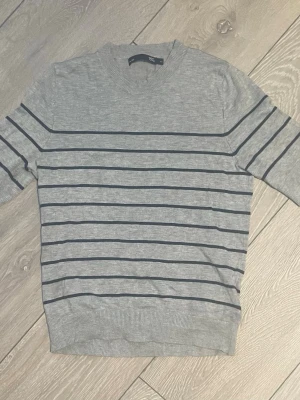 Grå stickad t-shirt - Snygg stickad t-shirt från Lager 157. Tröjan har rund halsringning och ribbade muddar vid ärmslut och nederkant. Använd fåtal gånger