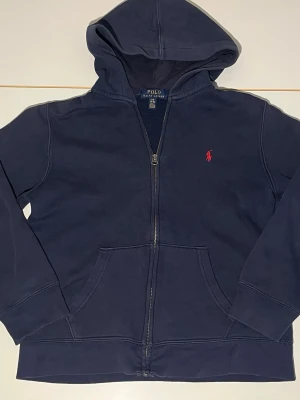 Mörkblå hoodie från Polo Ralph Lauren - Mörkblå hoodie från Polo Ralph Lauren med dragkedja, kängurufickor och klassisk röd logga broderad på bröstet. Tillverkad i mjuk bomullsblandning och har en stor huva. Perfekt för en avslappnad och stilren look.