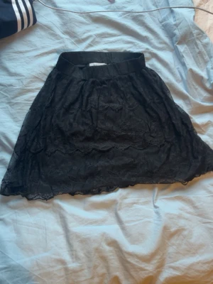 Svart spetskjol från Size & Needle - Supersöt svart kortkjol från Size & Needle i spetsmaterial. Kjolen har elastisk midja och ett fint spetsmönster över hela tyget. Perfekt att styla med en enkel topp eller hoodie för en cool look.