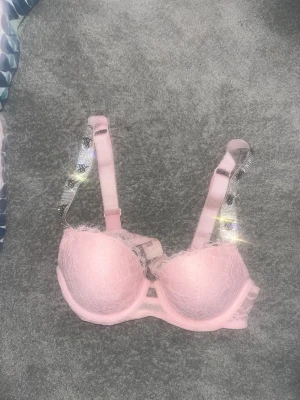 Rosa bh med spets och strass - Säljer en ljusrosa push-up bh med vadderade kupor och spetsdetaljer. Axelbanden är dekorerade med glittrande strass för en extra snygg touch. Perfekt för dig som vill ha något både sött och glamoröst. Köpte här på Plick men var för liten.