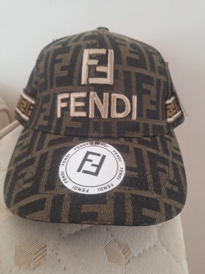 Fendi keps med logomönster - Snygg keps från Fendi med klassiskt FF-logomönster i svart och brunt. Stor broderad Fendi-logga framtill och detaljer på sidorna. Kepsen har böjd skärm och är tillverkad i ett robust tygmaterial. Perfekt för dig som vill sticka ut med en lyxig accessoar.