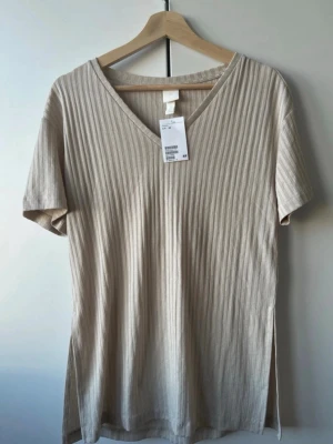 Beige t-shirt med v-ringning - Snygg beige t-shirt från H&M med ribbad struktur och v-ringad hals. Perfekt för en stilren och enkel look. Materialet är mjukt och bekvämt, vilket gör den lätt att bära hela dagen! Buda gärna💗
