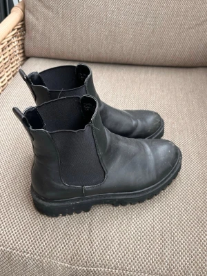 Svarta chelsea boots i skinn  - Svarta chelsea boots i läder med elastiska sidopaneler och grov, chunky sula. Klassisk siluett med dragflikar både fram och bak för enkel påtagning. Perfekta för dig som vill ha en stilren men edgy look. Passar till många olika outfits och ger en cool vibe.