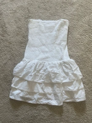 Vit axelbandslös klänning Gina Tricot - Söt vit axelbandslös klänning från Gina Tricot med smock upptill och volangkjol i två lager. Klänningen är kort och har broderade detaljer i kjolen som ger en romantisk känsla. Perfekt för varma dagar och sommarens alla tillfällen. Den har inte en ända fläck den är helt ny 