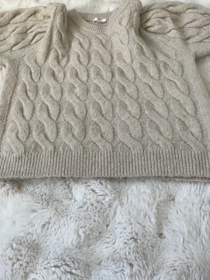 Beige kabelstickad tröja med puffärm - Trendig beige kabelstickad tröja med puffärmar och ribbade muddar. Tröjan har en rund halsringning och ett snyggt strukturmönster framtill. Perfekt för dig som gillar en stilren men ändå unik look.