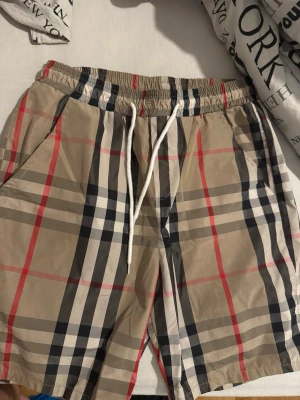 Beige rutiga shorts med snörning  - Snygga beige shorts med klassiskt rutigt mönster i svart, vitt och rött. Shortsen har elastisk midja med vit snörning och sidofickor. Perfekta för sommaren och ger en trendig look.