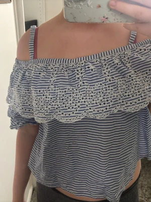 Randig offshoulder topp med spets - Söt blåvit randig offshoulder topp med volang och broderad spetsdetalj runt axlarna. Croppad modell med smala axelband och luftig passform, perfekt för varma dagar. Säljer då den inte passar mig