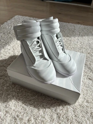 MaisonMargiela Future - Snygga vita high top sneakers med breda, vadderade remmar runt ankeln och klassisk snörning. Skorna har en stilren och futuristisk design med slät ovandel i syntetmaterial. Perfekta för dig som vill sticka ut med en clean look. 