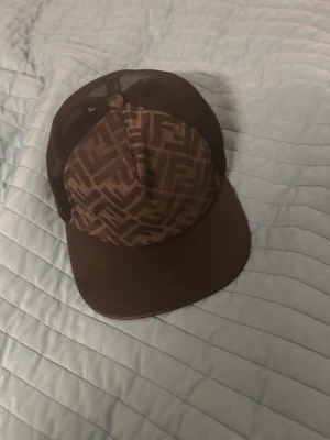 Brun keps med FF-mönster - Snygg brun keps från Fendi med klassiskt FF-mönster på framsidan och mesh-tyg baktill för extra ventilation. Kepsen har böjd skärm och justerbar passform. Perfekt accessoar för en trendig streetwear-look.