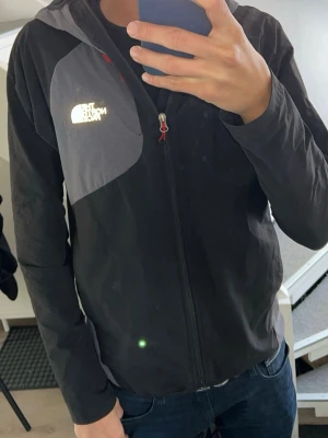 Svart vindjacka med reflex från The North Face - Snygg svart vindjacka från The North Face med grått parti över bröstet och vit logga. Jackan har dragkedja framtill, huva och är tillverkad i ett lätt, vindtätt material. Perfekt för blåsiga dagar och enkel att packa ner i väskan.