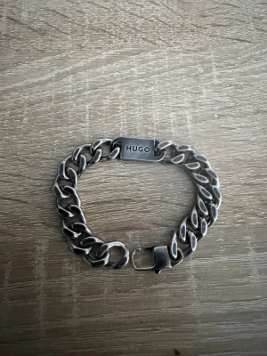 Silverfärgat kedjearmband från Hugo - Stilrent armband i silverfärgad metall från Hugo med breda kedjelänkar och en platta med ingraverad logga. Perfekt accessoar för en modern och edgy look.