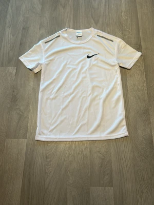 Vit Nike sport t-shirt - Vit sport t-shirt från Nike med svart swoosh-logga på bröstet och grå detaljer på axlarna. Tillverkad i ett lätt och ventilerande funktionsmaterial som passar perfekt för träning och aktivitet.