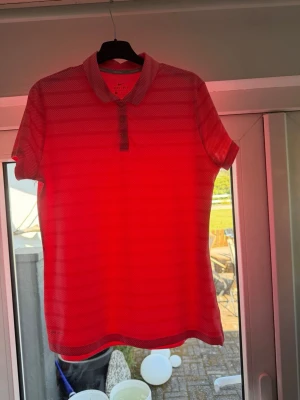 Röd Nike Golf pikétröja Dri-Fit - Snygg röd pikétröja från Nike Golf med Dri-Fit-material som andas. Tröjan har korta ärmar, krage och knappar framtill samt diskreta mörkröda ränder i mönstret. Perfekt för golf eller andra sportaktiviteter.