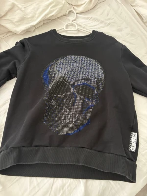 Svart sweatshirt med döskalle Philipp Plein - Cool svart sweatshirt från Philipp Plein med en stor glittrig döskalle framtill i silver och blått. Tröjan har rund hals, ribbade muddar och en metallogo i nacken. Perfekt för dig som gillar edgy streetstyle.
