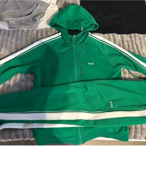 Grönt loungewear-set från BOSS - Snyggt grönt loungewear-set från BOSS med vit rand längs ärmar och ben. Setet består av en hoodie med dragkedja och matchande byxor, båda med BOSS-logga. Perfekt för chill dagar hemma. Mjukt material och sportig look.