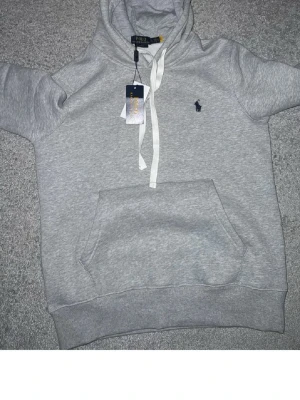 Grå hoodie från Polo Ralph Lauren - Snygg grå hoodie från Polo Ralph Lauren med klassisk huva och vita dragsnören. Har en stor magficka och den ikoniska lilla broderade loggan i mörkblått på bröstet. Perfekt för en chill och stilren look. Mjuk bomullsblandning som är skön att ha på sig. Finns även gråa snören som kommer med