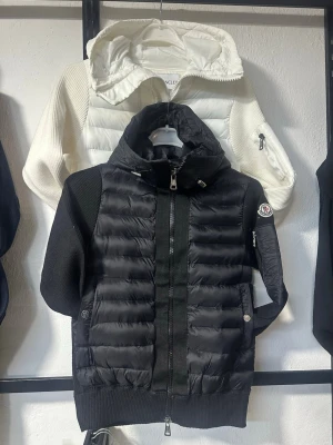 Svart puffer kardigan från Moncler - Snygg svart puffer kardigan från Moncler med quiltad front och huva. Jackan har stickade ärmar och sidor, dragkedja framtill och två fickor med tryckknapp. Moncler-logga på vänster ärm och justerbar huva. Perfekt för våren 