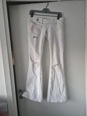 Vita utsvängda jeans med slitningar - Säljer ett par vita jeans med utsvängda ben och slitningar vid knäna och fickan. Jeansen har klassisk femficksdesign och låg midja. Dessa säljer jag för att de är för korta och jag vill köpa de som är "tall".