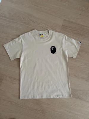 Beige BAPE t-shirt med logga - Säljer en beige t-shirt från BAPE med den klassiska svarta ap-loggan tryckt på bröstet. T-shirten har rund hals och korta ärmar, tillverkad i mjuk bomull för skön passform. Perfekt för dig som gillar streetwear och vill sticka ut.