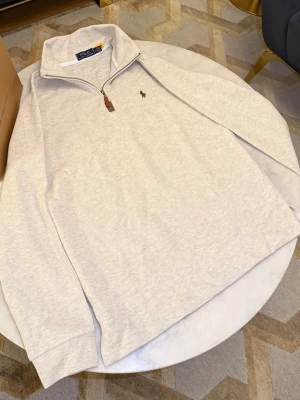 Ljusgrå halvzip från Polo Ralph Lauren - Ljusgrå stickad tröja från Polo Ralph Lauren med halv dragkedja och hög krage. Broderad brun logga på bröstet och ribbade muddar. Tröjan är i mjukt material och har en stilren, klassisk look som funkar året runt.