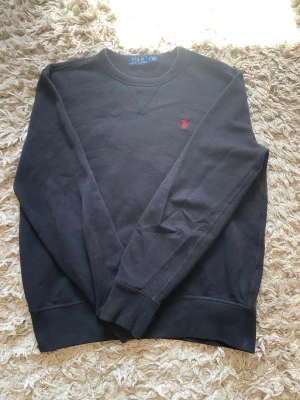 Svart sweatshirt från Polo Ralph Lauren - Svart crewneck sweatshirt från Polo Ralph Lauren med klassisk röd broderad logga på bröstet. Tröjan har rund halsringning, långa ärmar och ribbade muddar. Perfekt för en avslappnad och stilren look.