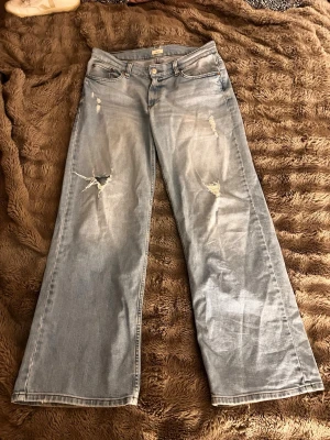 Ljusblåa utsvängda jeans med slitningar - Säljer ett par ljusblåa jeans med breda ben och coola slitningar framtill. Jeansen har klassisk femficksdesign och låg midja. Perfekta för en avslappnad och trendig look. Materialet är denim i bomull.