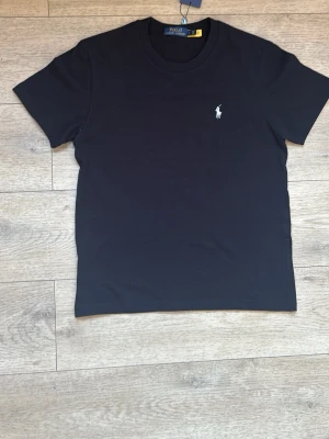 Svart t-shirt från Polo Ralph Lauren - Svart t-shirt från Polo Ralph Lauren med rund hals och korta ärmar. Den har en vit broderad polospelare på bröstet och är gjord i mjuk bomull. Enkel och stilren design som alltid funkar fick i födelsedagspresent men är för liten tyvärr