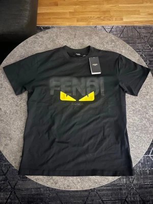 Svart Fendi t shirt - Cool svart t-shirt från Fendi med stora FENDI-bokstäver och ikoniska gula ögon på bröstet. Klassisk passform med rund hals och korta ärmar. Perfekt för dig som vill sticka ut med en lyxig streetstyle-look.