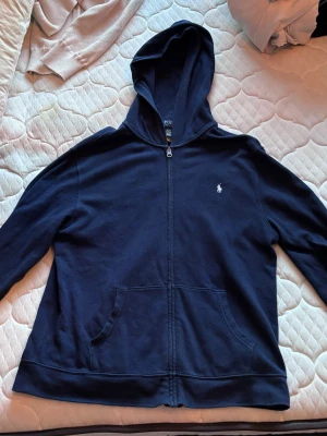 Mörkblå hoodie från Polo Ralph Lauren - Mörkblå hoodie med dragkedja från Polo Ralph Lauren. Klassisk design med huva, två fickor framtill och den vita logotypen broderad på bröstet. Storlek: XL (18-20)