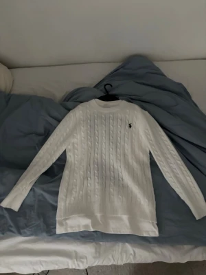 Vit kabelstickad tröja från Ralph Lauren - Vit kabelstickad tröja från Ralph Lauren med rund halsringning och lång ärm. Tröjan har det klassiska broderade logotypen på bröstet och är tillverkad i mjukt material som passar perfekt för kyligare dagar. Passar xs