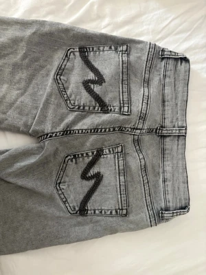 Gråa bootcutjeans från Nelly - Säljer ett par gråa bootcutjeans från Nelly med svarta zigzag-detaljer på bakfickorna. Jeansen har låg midja och klassisk femficksdesign. Perfekta för dig som gillar en avslappnad och trendig stil.