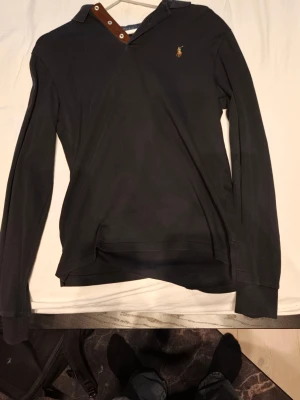 Svart långärmad henley från Polo Ralph Lauren - Svart långärmad henley från Polo Ralph Lauren med klassisk knappslå i brunt och silverdetaljer. Skjortan har en broderad logga på bröstet och är tillverkad i mjuk bomull. Perfekt för en stilren och avslappnad look.