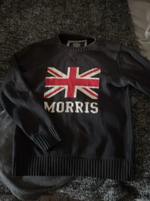 Mörkblå stickad tröja Morris - Mörkgrå stickad tröja från Morris med Union Jack-flagga och MORRIS-text framtill. Tröjan har ribbstickade muddar vid ärmslut, hals och nederkant. Perfekt för dig som gillar klassisk brittisk stil och vill ha något som sticker ut.