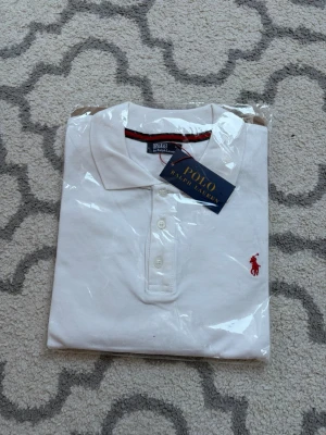 Vit pikétröja från Polo Ralph Lauren - (Helt ny)Klassisk vit pikétröja från Polo Ralph Lauren med röd broderad logga på bröstet. Tröjan har krage, knappar framtill och är tillverkad i stretchigt bomullsmaterial. Perfekt för en stilren och sportig look.