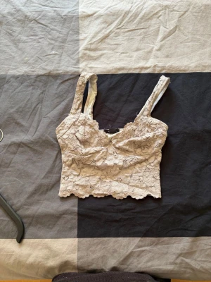 Beige spetsbody med breda axelband - Snygg beige body i spets med breda axelband och blommigt mönster. Modellen är croppad och har en mjuk insida för extra komfort. Perfekt för att skapa en romantisk look med en modern twist.
