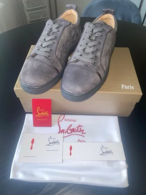 Gråa sneakers från Christian Louboutin - Snygga gråa sneakers från Christian Louboutin i mocka med klassisk röd sula och diskret logga på sidan. Skorna har rund tå, gråa skosnören och låg profil. Perfekta för dig som vill ha en lyxig och stilren look.