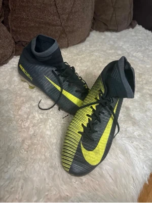 Nike Mercurial fotbollsskor svart/gul - Säljer ett par Nike Mercurial fotbollsskor i svart med gula detaljer och högt skaft. Skorna har snörning och en tydlig Nike-logga på sidan. Perfekta för dig som vill ha bra grepp och stil på planen.