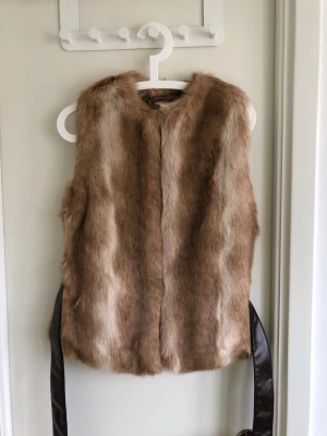 Beige pälsväst med brunt band - Snygg beige pälsväst i fuskpäls med mjukt material och brunt satinband i midjan. Västen är ärmlös och har en rak passform, perfekt för att lyfta din outfit med en trendig touch.