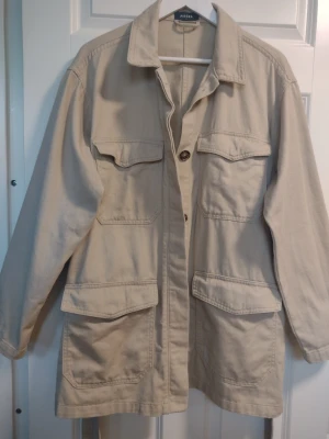 Beige jacka/jeansjacka/sommarjacka/utilityjacka/overshirt från Pieces - Beige jacka i 100% bomull. Avtagbart knytband i midjan. Stängs med bruna fina knappar och fickorna med tryckknappar. Sparsamt använd och i fint skick. Storlek XL. Även fin som oversize.