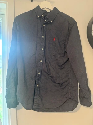Mörkblå skjorta från Ralph Lauren - Klassisk mörkblå skjorta från Ralph Lauren med button-down krage och röd broderad logga på bröstet. Skjortan har långa ärmar, vita knappar och är tillverkad i bomull. Perfekt för en stilren och avslappnad look.