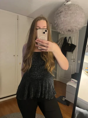 Glittrig svart ärmlös topp - Snygg svart topp med glittrigt tyg och volang nedtill. Ärmlös modell med figurnära passform och lätt utsvängd nederdel. Perfekt för dig som vill sticka ut lite extra. 