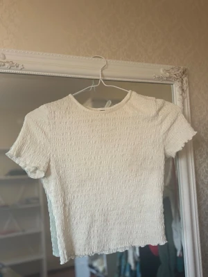 Vit ribbad topp från H&M - Säljer en vit ribbad topp från H&M med korta ärmar och rund hals. Toppen har en smockad struktur och är lätt croppad med vågig kant nertill. Perfekt att styla med jeans eller kjol för en enkel och fräsch look. Tyvärr har tröjan en liten rosa fläck, som man kan se på en av bilderna.