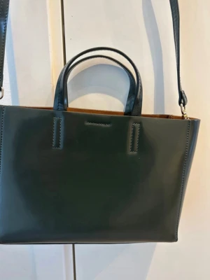 Mörkgrön handväska från Wera - Stilren mörkgrön handväska från Wera i glansigt skinn. Väskan har både handtag och en lång axelrem för olika bärsätt. Rektangulär form med minimalistisk design och diskret logga framtill. Perfekt för dig som gillar klassiska och trendiga accessoarer. Fick den som present men den är oanvänd.