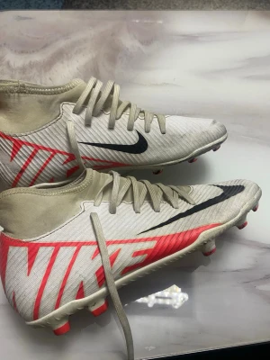 Nike vita och röda fotbollsskor - Snygga fotbollsskor från Nike i vitt med röda detaljer och svart swoosh. Skorna har högt skaft, snörning och mönstrad sula för bra grepp på planen. Tillverkade i syntetmaterial och designade för att ge bra stöd och känsla vid spel. Inte mycket använda