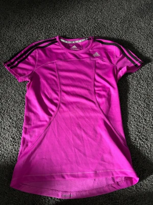 Rosa tränings-tshirt från Adidas - Snygg rosa tränings-tshirt från Adidas med svarta ränder på axlarna och logga på bröstet. T-shirten har figurnära passform, korta ärmar och är tillverkad i ett lätt och ventilerande funktionsmaterial som passar perfekt till träning.