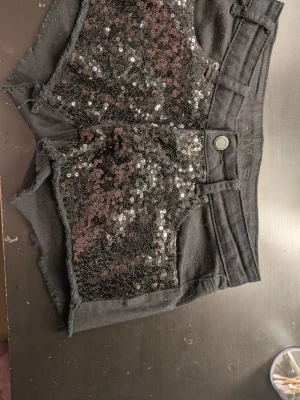 Svarta paljettshorts - Säljer ett par svarta jeans-shorts då den inte används längre. Det sitter svarta paljetter framtill och är vanliga baktill. Dom e även normal höga i midjan💞