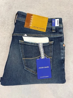 Blå jeans från Jacob Cohën - Snygga blå jeans från Jacob Cohën med klassisk femficksdesign och raka ben. Jeansen har en gulbrun patch med logga bak och subtila slitningar framtill. Tillverkade i mjuk denim med en modern tvättad look. Perfekta för en trendig och avslappnad stil.