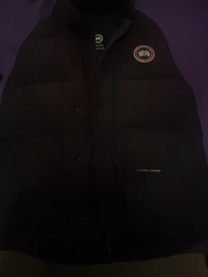 Svart dunväst från Canada Goose - Säljer en svart dunväst från Canada Goose med klassisk logotyp på bröstet och diskret varumärkesbrodyr. Västen har hög krage och är fylld med dun för extra värme. Perfekt för lager-på-lager och riktigt snygg till streetwear-stilen.